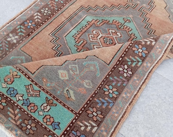 Puede incluir: Una alfombra rectangular plegada con un patrón geométrico detallado. La alfombra presenta un diseño central en tonos melocotón, gris y turquesa, enmarcado por un borde marrón con motivos florales. El diseño de la alfombra es intrincado y tradicional.
