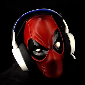 May include: A red and black Deadpool mask with white headphones on top. The mask has a black eye mask with white eyes.
