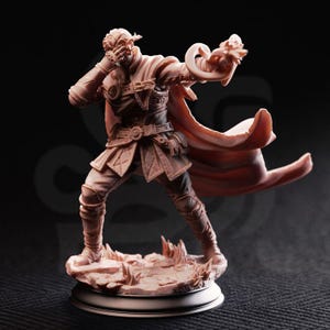 May include: A detailed figurine of a character in a dynamic pose, wearing a detailed outfit with a flowing cape. The figure is in a fighting stance, with one hand raised to the face and the other holding a curved object. The base is a textured, rocky terrain.