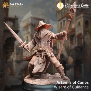 Puede incluir: Una figura miniatura pintada de un mago sosteniendo un bastón y una espada. El mago lleva una túnica larga y un sombrero puntiagudo. La figura está de pie sobre una base rocosa. El texto "Artemis of Canos Wizard of Guidance" está impreso debajo de la figura.