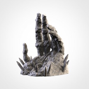Può includere: Una scultura di mano in pietra grigia, con bordi taglienti e irregolari, che si erge verso l'alto. La mano è circondata da pietre appuntite più piccole.