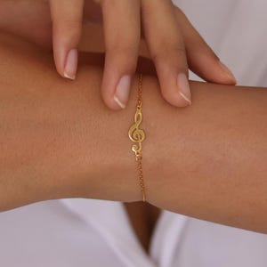 May include: A gold chain bracelet with a treble clef charm. The bracelet is on a person's wrist.
