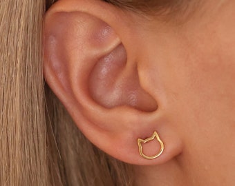 May include: A close-up of a person's ear with a small gold cat-shaped earring. The earring is a simple outline of a cat's head.