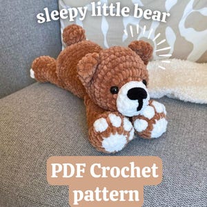 Könnte beinhalten: Ein brauner gehäkelter Bär mit weißen Akzenten an Pfoten und Schnauze. Der Bär liegt mit ausgestreckten Armen da. Der Text auf dem Bild lautet "sleepy little bear" und "PDF Crochet pattern".