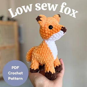 Könnte beinhalten: Ein gehäkelter Fuchskuscheltier in Orange-, Weiß- und Brauntönen. Der Fuchs hat schwarze Augen und trägt die Aufschrift "Low sew fox" und "PDF Crochet Pattern". Das Plüschtier wird in einer Hand gehalten.