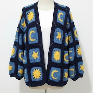 May include: A navy blue, open-front crochet cardigan featuring a repeating pattern of yellow suns, moons, and stars on blue squares. The sleeves are three-quarter length, and the garment is displayed on a mannequin.