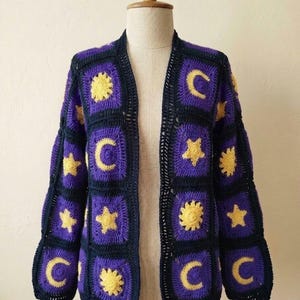 May include: A handmade, open-front cardigan in a celestial theme. The crochet design features square panels in purple, black, and yellow. The squares display suns, moons, and stars. The cardigan is displayed on a mannequin.