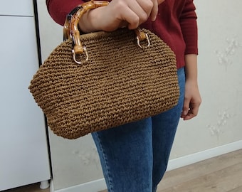 May include: A brown woven handbag with a bamboo handle and gold-colored metal accents. The bag has a rounded shape and is held by a person wearing a burgundy sweater and blue jeans. The bag's texture is visible.