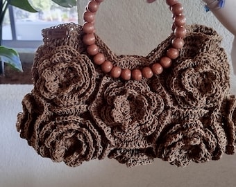 May include: A brown crocheted handbag with a wooden beaded handle. The bag features a floral design with multiple crocheted roses. The handle is made of round, light brown wooden beads. The bag is a unique, handmade accessory.