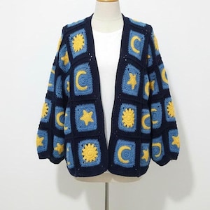 May include: A navy blue crochet cardigan featuring a celestial design. The open-front sweater has square panels with blue backgrounds and yellow sun, moon, and star motifs. The sleeves are three-quarter length.