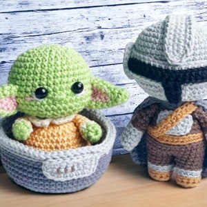May include: A green crocheted baby Yoda doll sitting in a gray crocheted pod, next to a gray and brown crocheted Mandalorian doll.