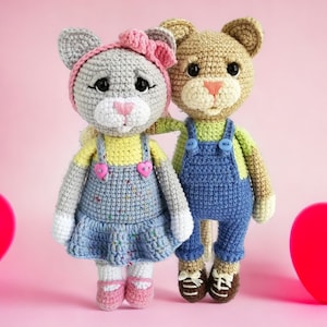 Może przedstawiać: Szary szydełkowy kot z różową kokardką i niebiesko-żółtym strojem trzyma brązowego szydełkowego kota w zielonej koszuli i niebieskich ogrodniczkach. Oba koty stoją na różowym tle.