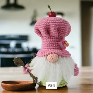 May include: A pink crocheted gnome wearing a pink and white chef's hat with a cherry on top. The gnome has a white beard and is holding a spoon. The gnome is sitting on a wooden table with a wooden bowl in front of it. The gnome is labelled #54.