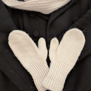 Könnte beinhalten: Ein Paar weiße Strickhandschuhe mit gerippter Struktur. Die Handschuhe sind zusammengefaltet und auf einem schwarzen Mantel gelegt.