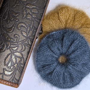 Könnte beinhalten: Eine verzierte, rechteckige Box mit einem floralen Muster in Gold und Schwarz, neben zwei gestrickten, kreisförmigen Gegenständen. Einer ist in einem gedämpften Goldton gehalten, der andere in Staubblau. Die Artikel scheinen Hüte oder Dekorationen zu sein.