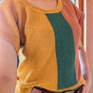Könnte beinhalten: Ein kurzärmeliges Strickoberteil mit einem Color-Block-Design aus vertikalen Bahnen in Senfgelb, Waldgrün und Terrakotta-Orange. Das Kleidungsstück hat einen Rundhalsausschnitt mit geripptem Kragen und passenden gerippten Abschlüssen an den Ärmeln.
