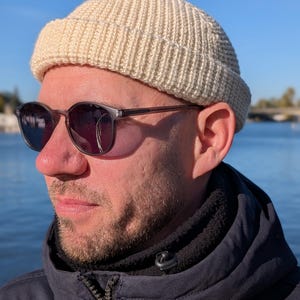 Può includere: Un berretto lavorato a maglia a coste color crema. Il cappello è indossato con occhiali da sole con montatura scura e una giacca blu navy. Lo sfondo mostra un corpo idrico e un cielo azzurro.