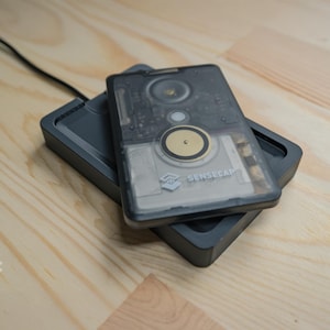 Peut inclure: Un chargeur noir avec un appareil rectangulaire transparent gris sur le dessus. L'appareil a un bouton circulaire avec un anneau doré et le texte "Sensecap" imprimé dessus.