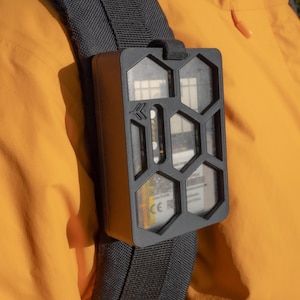 Może przedstawiać: Czarne, prostokątne urządzenie z wzorem plastra miodu i przezroczystym okienkiem z tworzywa sztucznego. Urządzenie jest przymocowane do czarnego paska i noszone na ramieniu osoby.