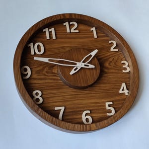 Op de afbeelding: Een ronde houten wandklok met een donkerbruine afwerking. De klok heeft grote, lichtgekleurde cijfers en wijzers. De wijzerplaat heeft een cirkelvormig ontwerp met een verhoogd midden en een gladde, gepolijste afwerking. Diameter ca. 30 cm.
