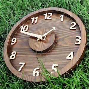 May include: A round wooden clock with a dark brown finish. The clock face features large, light-colored numerals and hands shaped like a saw and a hammer. The clock is set against a background of green grass.