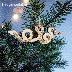 May include: A wooden Christmas ornament shaped like a snake with intricate details. The ornament is light brown and has a string for hanging.