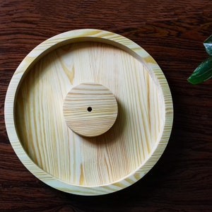 Puede incluir: Una bandeja redonda de madera con un disco redondo de madera más pequeño en el centro. La bandeja tiene un acabado de madera natural y es perfecta para servir aperitivos o exhibir objetos pequeños.
