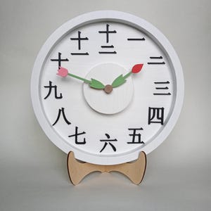 May include: White wall clock with black Japanese numerals. The clock face has a white background with a small white circle in the center. The clock hands are green and the tips are red and pink. The clock is on a wooden stand.