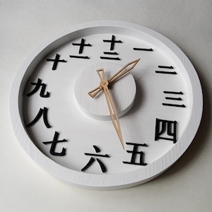 May include: White round wall clock with black Japanese numerals and wooden clock hands. The clock face has a white background with a raised white center. The clock hands are made of light brown wood.