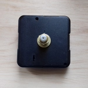 May include: A black square clock movement with a brass-colored nut and a white plastic piece in the center. The clock movement has a small hole at the top and a rectangular slot at the bottom. The background is a light wood grain.