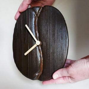 Op de afbeelding: Een donkerbruine houten klok met gouden wijzers. De wijzerplaat van de klok is ovaalvormig en heeft een houtnerfpatroon.