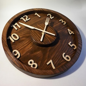 Könnte beinhalten: Eine dunkelbraune Wanduhr aus Holz mit weißen Zahlen und einem Gabel- und Messerdesign in der Mitte. Die Uhrzeiger sind golden.