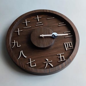 Pode incluir: Um relógio de madeira redondo com numerais chineses e um símbolo yin-yang. O mostrador do relógio é castanho escuro com numerais mais claros. Os ponteiros são de cor clara e o símbolo yin-yang é preto e branco.