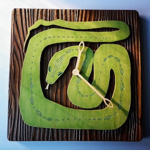 May include: A wooden clock with a green snake design. The snake is coiled around the clock face and has a detailed pattern of leaves and dots. The clock hands are gold.