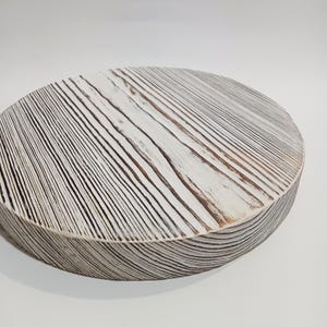 May include: A round, wooden decorative piece with a white-washed finish. The surface features a series of parallel, dark lines, creating a textured, striped pattern. The edges of the piece are visible, showing the layered construction.