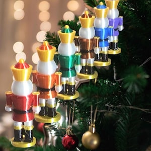 Pode incluir: Um conjunto de seis enfeites de quebra-nozes coloridos pendurados em uma árvore de Natal. Os enfeites são feitos de plástico impresso em 3D e apresentam uma variedade de cores, incluindo vermelho, laranja, verde, marrom, azul e roxo.