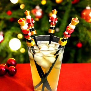 May include: Four black and gold cocktail stirrers with red and white nutcracker figurines on top. The stirrers are in a glass with ice and a light yellow liquid.