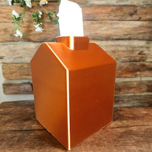May include: A terracotta-colored tissue box dispenser shaped like a house. The dispenser has a white trim and a white tissue box inside.