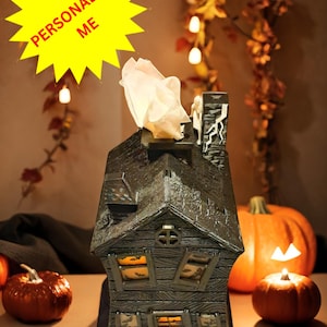 May include: A black and gray haunted house shaped tissue box holder with a lit window. The holder is sitting on a table with two orange pumpkins and a lit candle.
