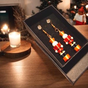 Può includere: Un paio di orecchini a forma di schiaccianoci dorati in una scatola regalo nera. Gli orecchini sono realizzati con perline rosse, bianche e dorate e presentano un design a forma di schiaccianoci.