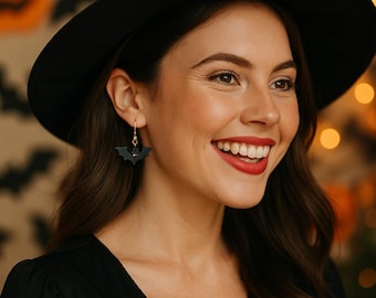 May include: A woman wearing a black witch hat and black dress smiles, showcasing bat-shaped earrings. The earrings are black with gold-colored hooks. The background features Halloween decorations, including pumpkin and bat silhouettes.