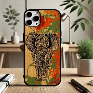 Puede incluir: Una funda de teléfono negra con un patrón de veta de madera y una ilustración detallada de un elefante con un diseño de mandala. El elefante es blanco y negro y el fondo es verde, naranja y marrón. La funda tiene un borde negro y la palabra "utopia" está impresa en la parte inferior.