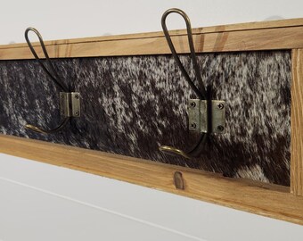 May include: A wooden coat rack with a cowhide backing and four metal hooks. The hooks are a dark bronze color and have a simple design.