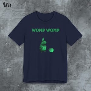 Può includere: Maglietta blu navy con una creatura verde dei cartoni animati che indossa un cappello da festa e tiene una palla verde. Il testo "WOMP WOMP" è stampato in verde sulla maglietta.