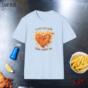 Puede incluir: Camiseta azul claro con un gráfico de un corazón hecho de patatas fritas y pescado. El texto en la camiseta dice "I love you more than a chippy tea."