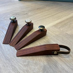 Puede incluir: Cuatro topes de puerta de madera con pomos decorativos. Tres tienen pomos en la parte superior, uno con un lazo de cuero. Los topes de puerta son de madera marrón oscuro y tienen forma de cuña. Los pomos tienen diferentes diseños.