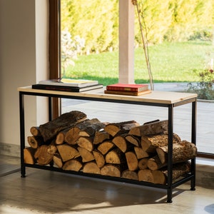 May include: A black metal wood rack with a white wooden top. The rack is filled with stacked firewood logs. Two books are on top of the rack. The rack is in front of a window with a view of a green lawn and trees.