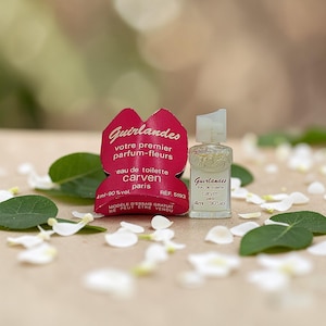 May include: A miniature perfume bottle and a red paper label with French text. The bottle is clear with a white cap and contains a light-coloured liquid. The label reads "Guirlandes votre premier parfum-fleurs eau de toilette carven paris 4ml-90% vol."