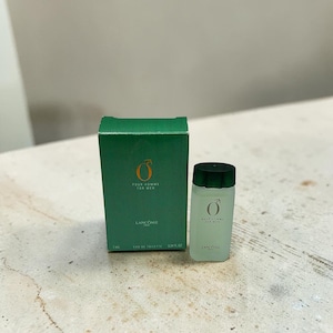 Może przedstawiać: Zielone pudełko i mała butelka perfum. Na pudełku i butelce widnieje napis "Pour Homme For Men" i "Lancôme". Butelka ma ciemnozieloną zakrętkę i zawiera przezroczysty płyn. Na pudełku widnieje napis "Eau de Toilette 7ml".