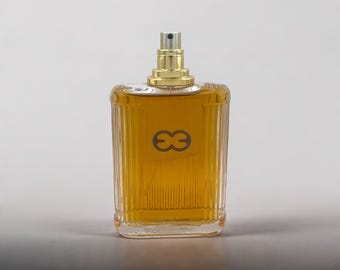 May include: A rectangular glass perfume bottle with vertical ribbed detailing containing amber-colored liquid. The bottle features a gold-toned spray nozzle and a dark brown logo consisting of two interlocking shapes on the front surface.